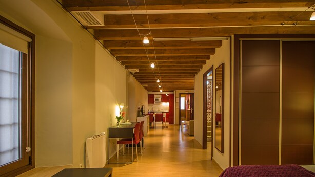 Una habitación de hotel con piso de madera, una cama con manta morada, un área de comedor con mesa y sillas, y una cocina al fondo.
