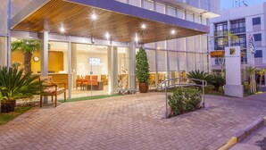 Entrada de hotel moderna com área de estar externa coberta, balcão de recepção e um grande vaso de plantas.