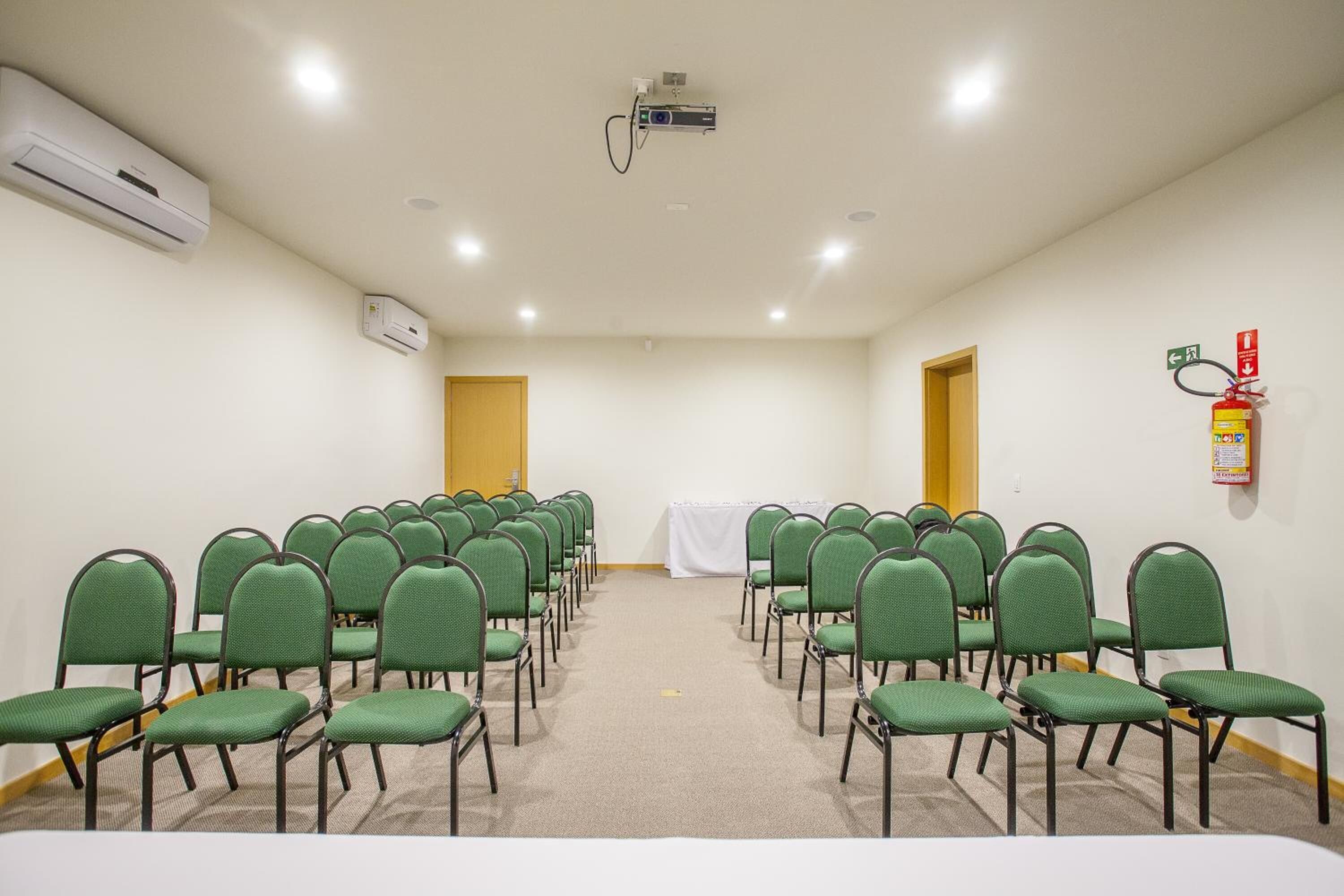 Uma sala de conferências com fileiras de cadeiras verdes, um projetor, aparelhos de ar condicionado e um extintor de incêndio.