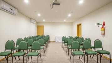 Uma sala de conferências com fileiras de cadeiras verdes, um projetor, aparelhos de ar condicionado e um extintor de incêndio.