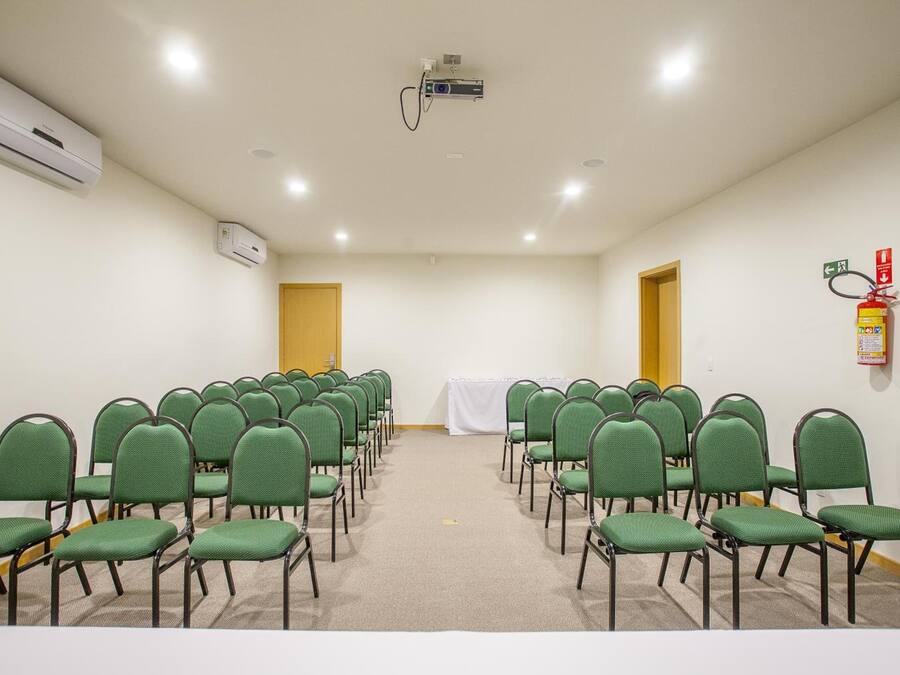 Uma sala de conferências com fileiras de cadeiras verdes, um projetor, aparelhos de ar condicionado e um extintor de incêndio.