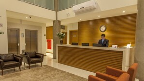 Uma área de recepção de hotel com um balcão de madeira, um recepcionista, duas poltronas de couro e piso coberto de carpete.