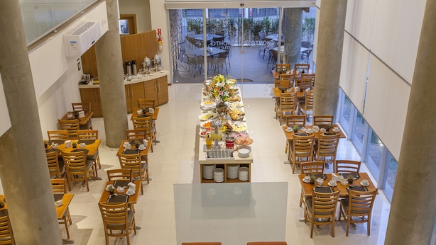 Ampla área de jantar de hotel, com várias mesas e cadeiras, um arranjo floral central e vista para o pátio exterior através de grandes janelas.