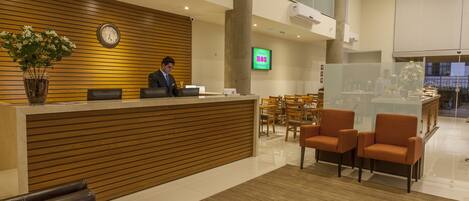 A hotel reception area with a wooden reception desk, a clock, and a TV screen.
