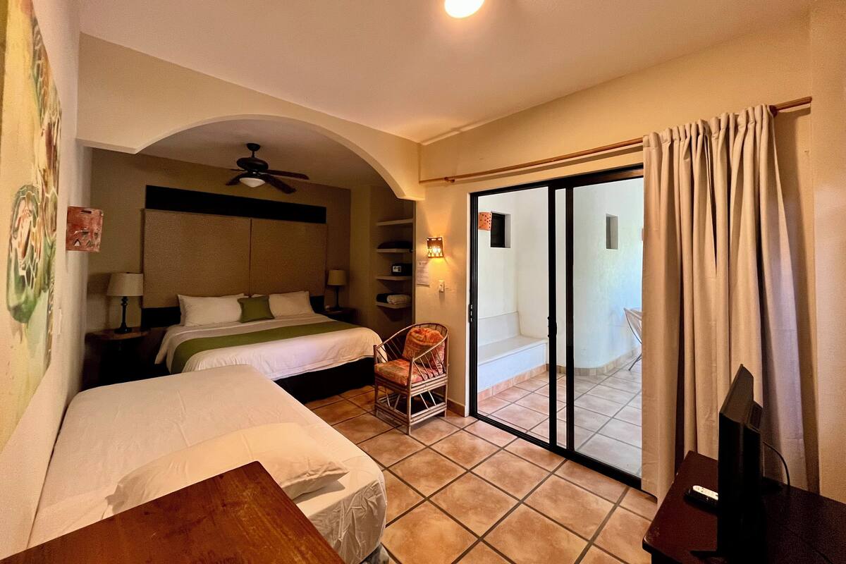 Habitación de hotel con dos camas, ventilador de techo, puerta corrediza al balcón y un televisor sobre una mesita.