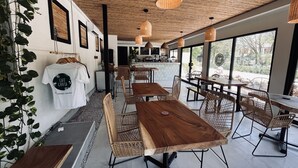 Una cafetería con mesas y sillas de madera, una zona de bar y grandes ventanales con vistas al exterior.