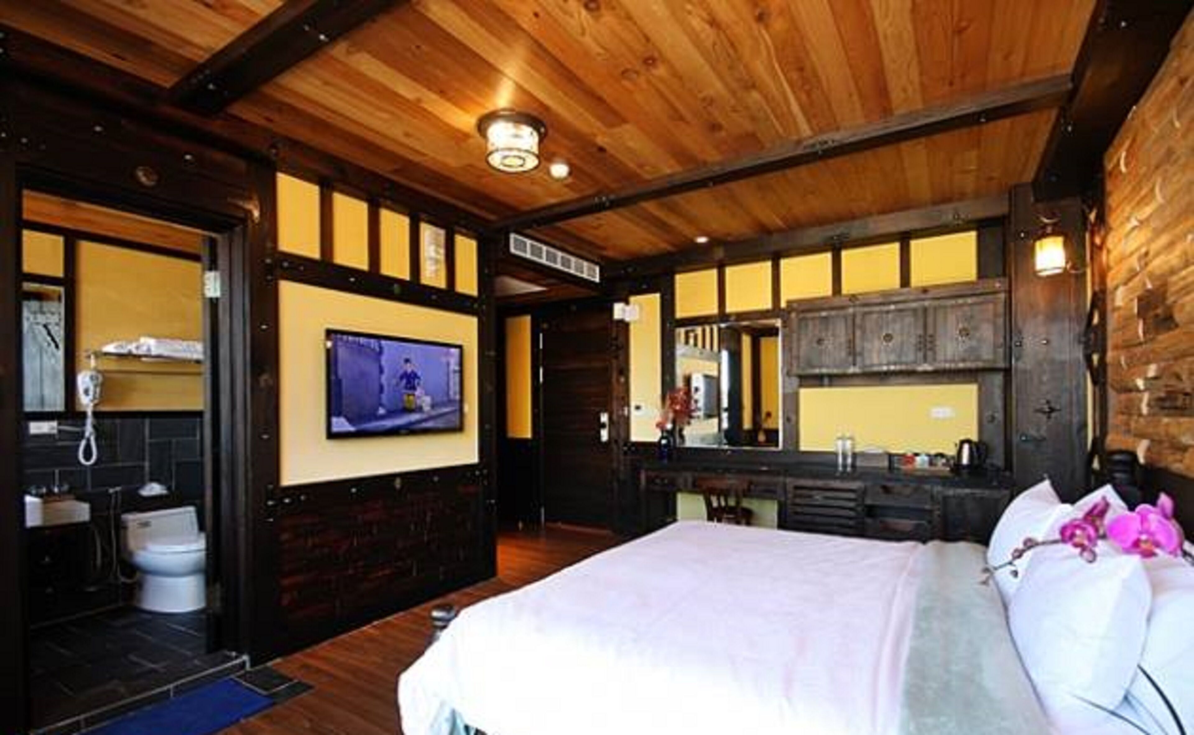 A room with a wooden ceiling, a television mounted on the wall, a bed with white bedding, a bathroom with a toilet and sink, and a mirror above the sink.