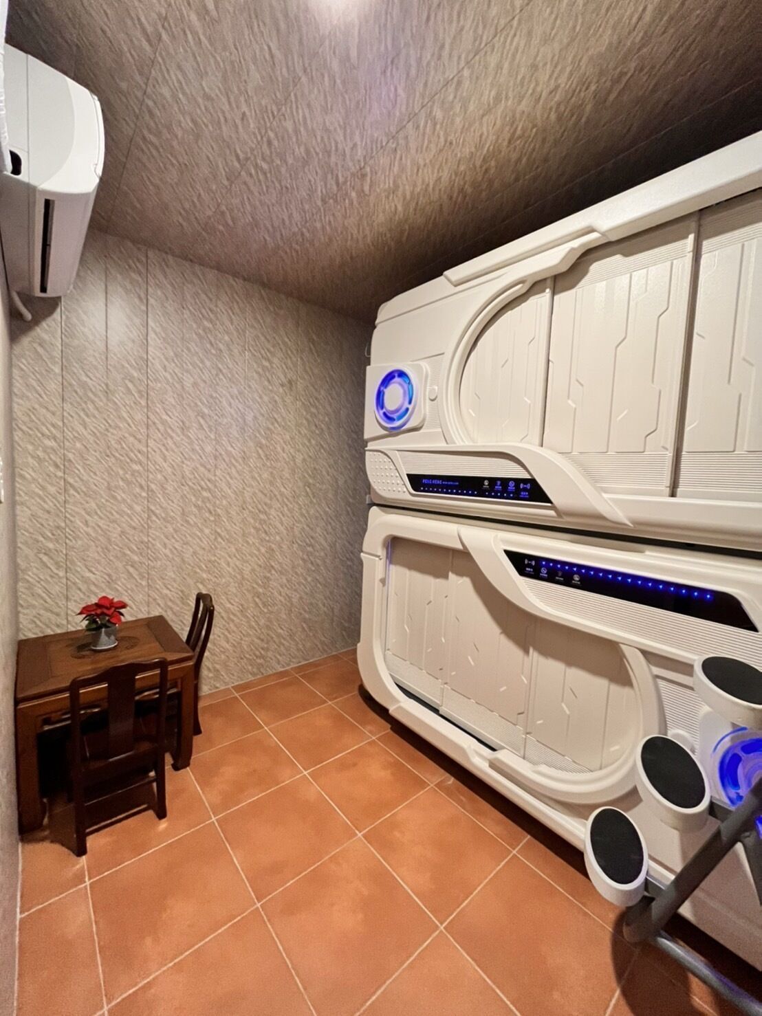 A room with a massage chair, a small dining table with chairs, and a wall-mounted air conditioner.