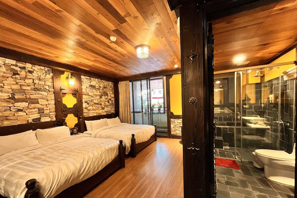 Habitación con dos camas, pared de piedra, techo de madera y baño con cabina de ducha de vidrio.