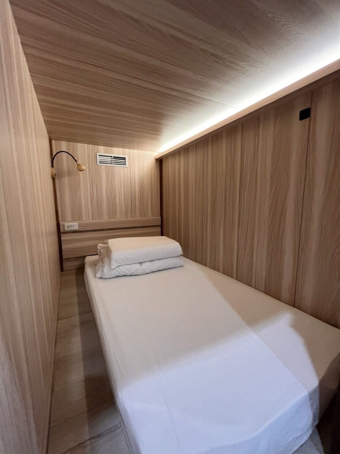 A narrow, single-bed room with wooden paneling on the walls and ceiling, a bedside table with a lamp, and a vent on the wall.