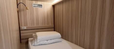 A narrow, single-bed room with wooden paneling on the walls and ceiling, a bedside table with a lamp, and a vent on the wall.