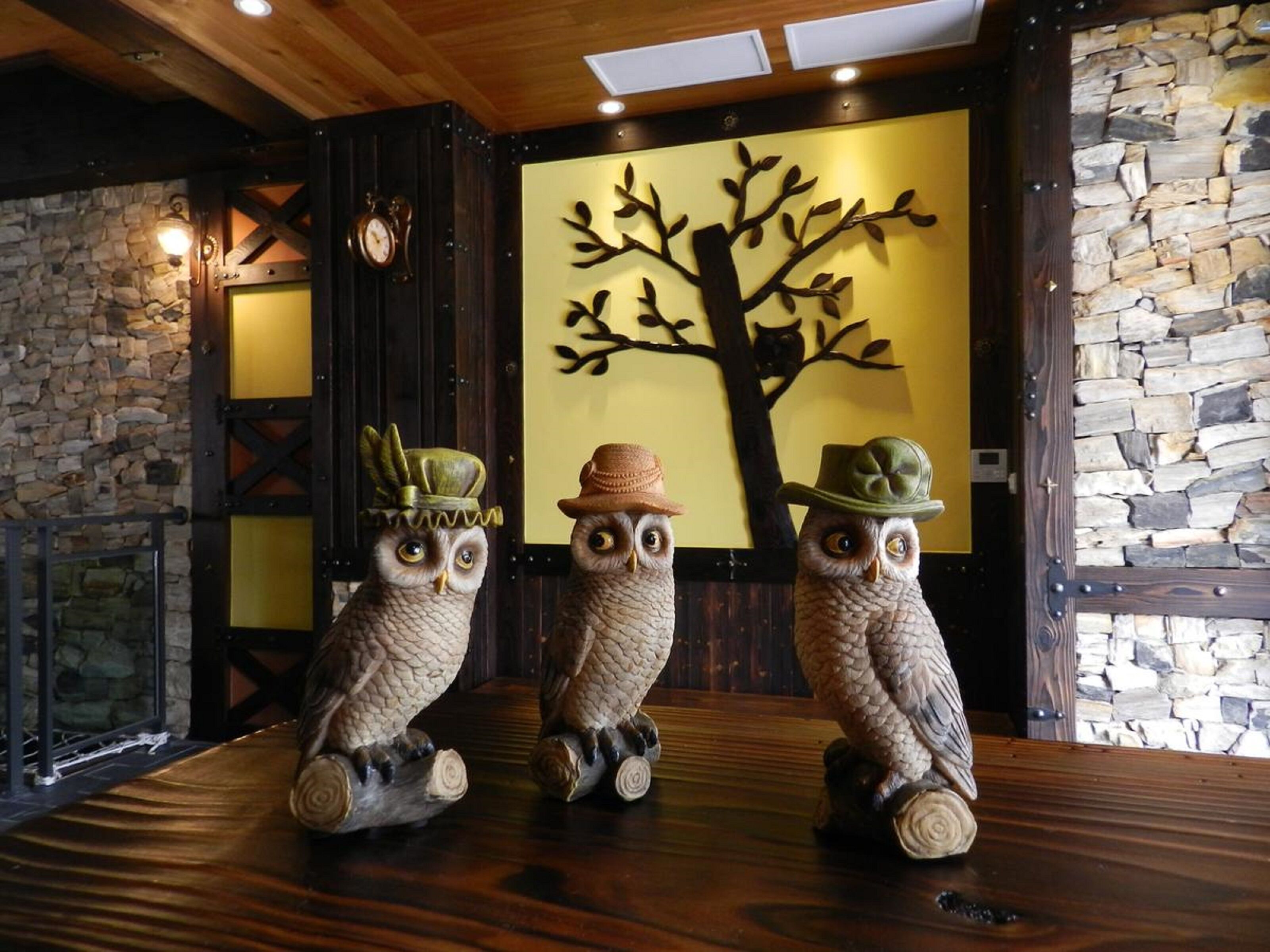 Tres estatuas de búhos sobre una mesa de madera, con una pared de piedra y un mural de un árbol al fondo.
