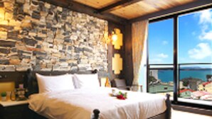A bedroom with a stone wall, a large bed, a nightstand, and a view of the sea through a large window.