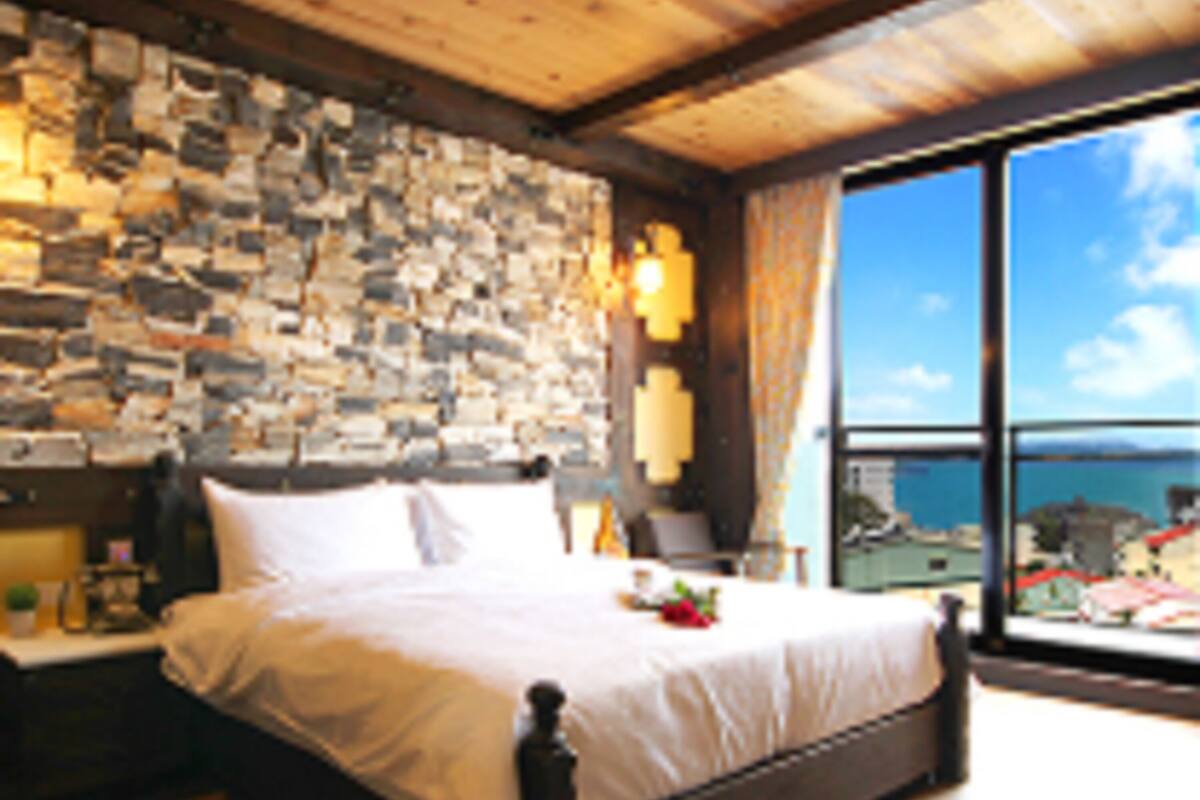 Un dormitorio con pared de piedra, una cama grande, una mesita de noche y vista al mar a través de un ventanal amplio.