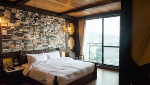 A bedroom with a stone wall, a wooden ceiling, a large window with a view, and a bed with white linens.