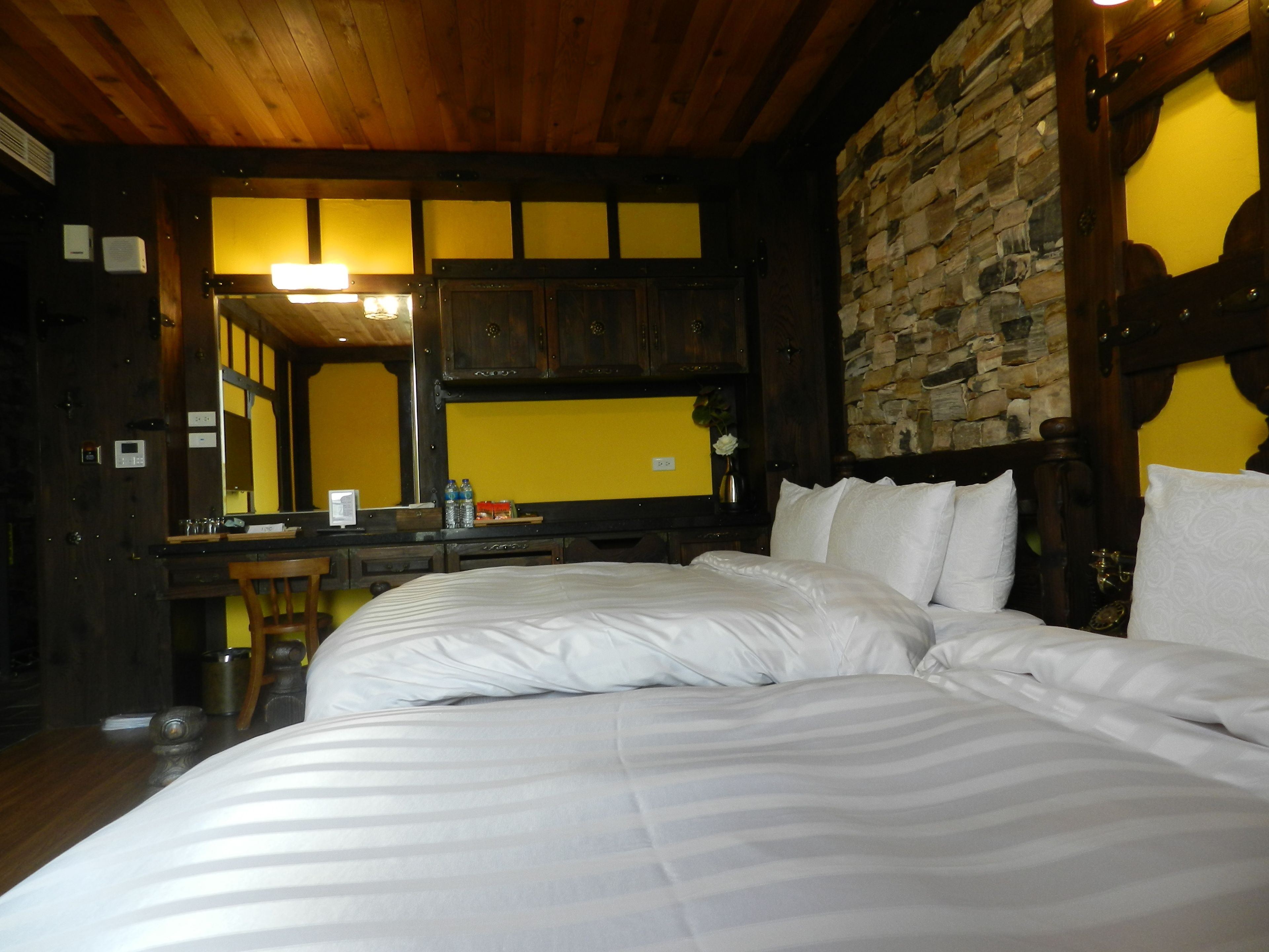 A hotel room with a large bed, wooden ceiling, stone wall, and a wooden desk with a mirror.
