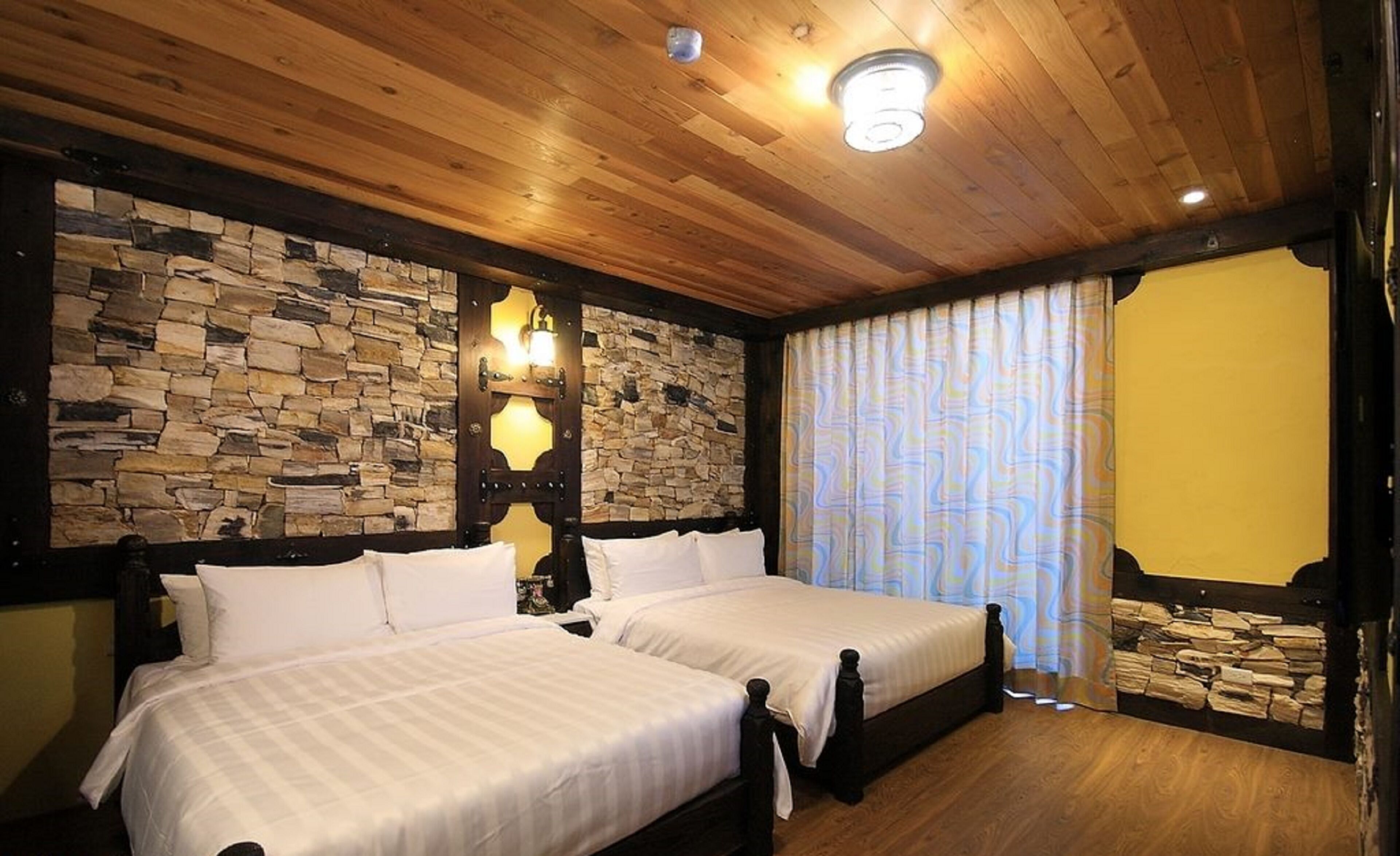 Una habitación con dos camas, paredes de piedra, techo de madera y un amplio ventanal con cortinas estampadas.