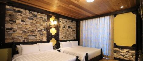 Una habitación con dos camas, paredes de piedra, techo de madera y un amplio ventanal con cortinas estampadas.