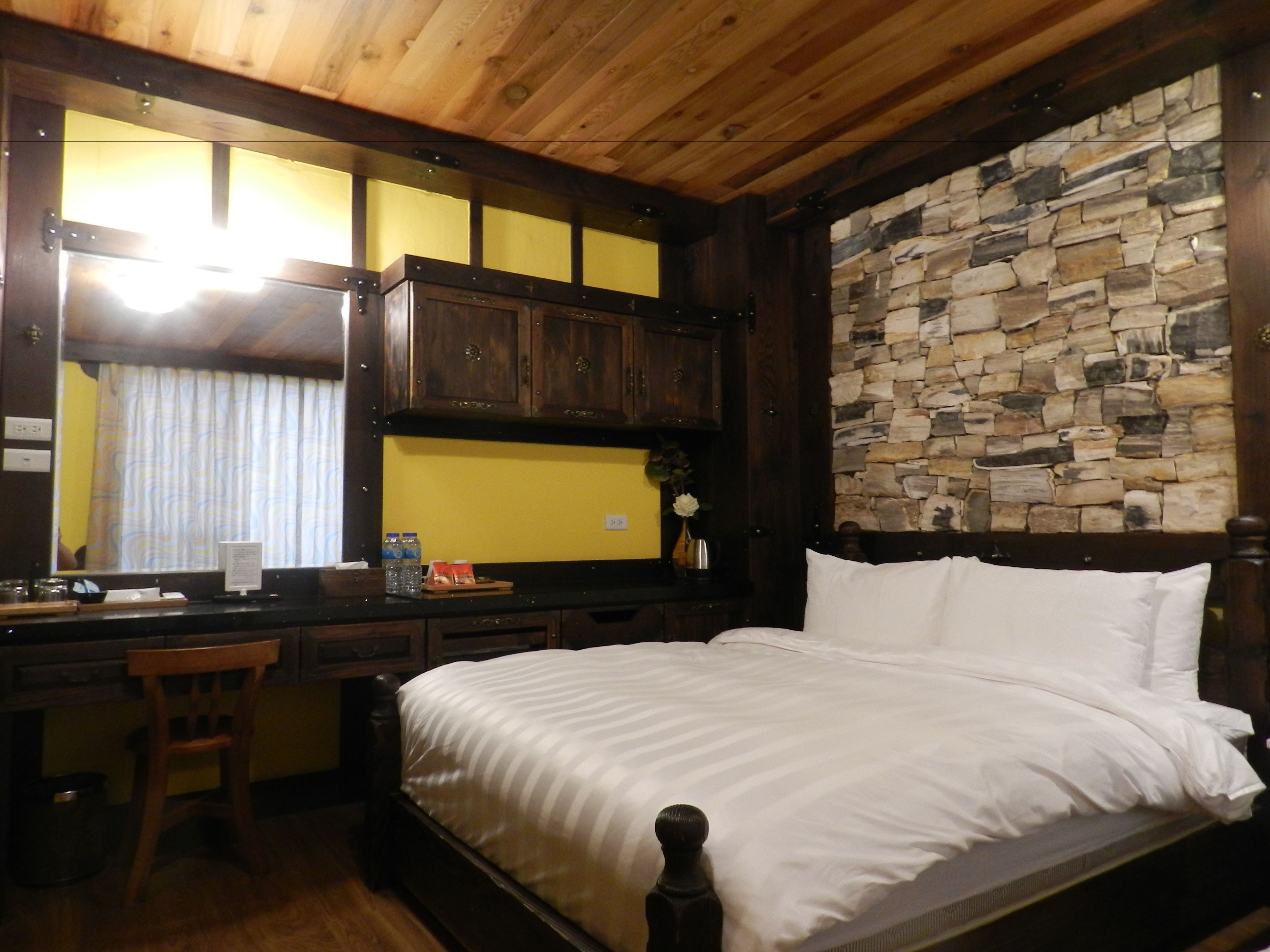Un dormitorio con techo de madera, pared de piedra, cama con ropa de cama blanca, escritorio con silla y ventana con cortinas.