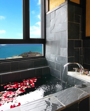 Une salle de bain dotée d’une grande fenêtre, d’une baignoire remplie de pétales de roses et offrant une vue sur la mer.