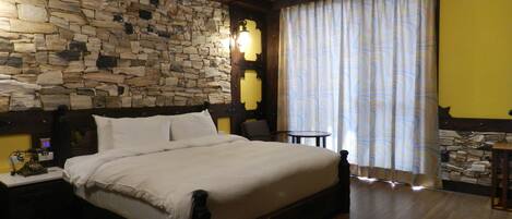 A bedroom with a stone wall, wooden ceiling beams, a bed with white linens, a nightstand, a small table, and a window with curtains.