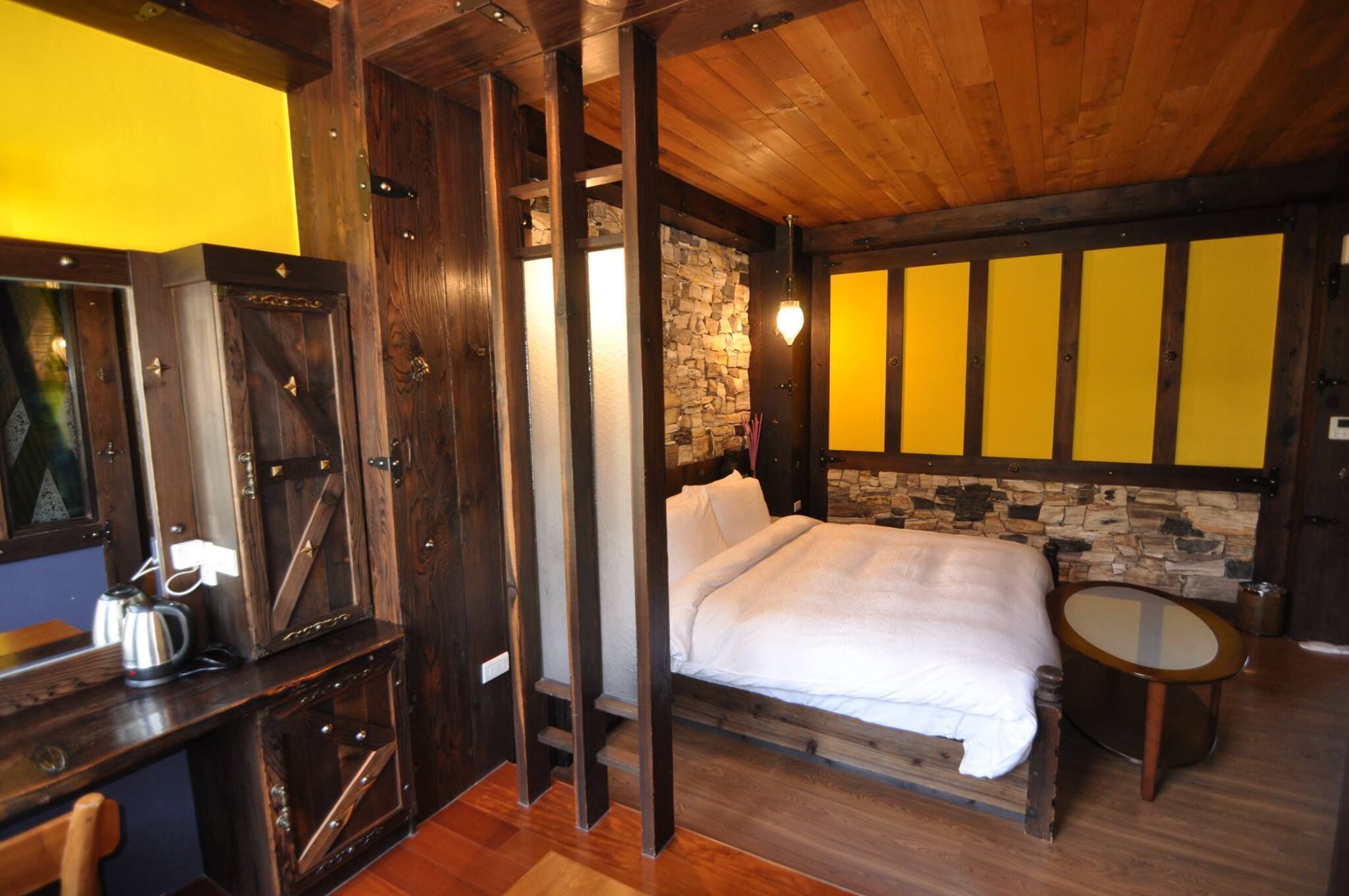 A room with a bed, a small table, a mirror, and a stone wall.