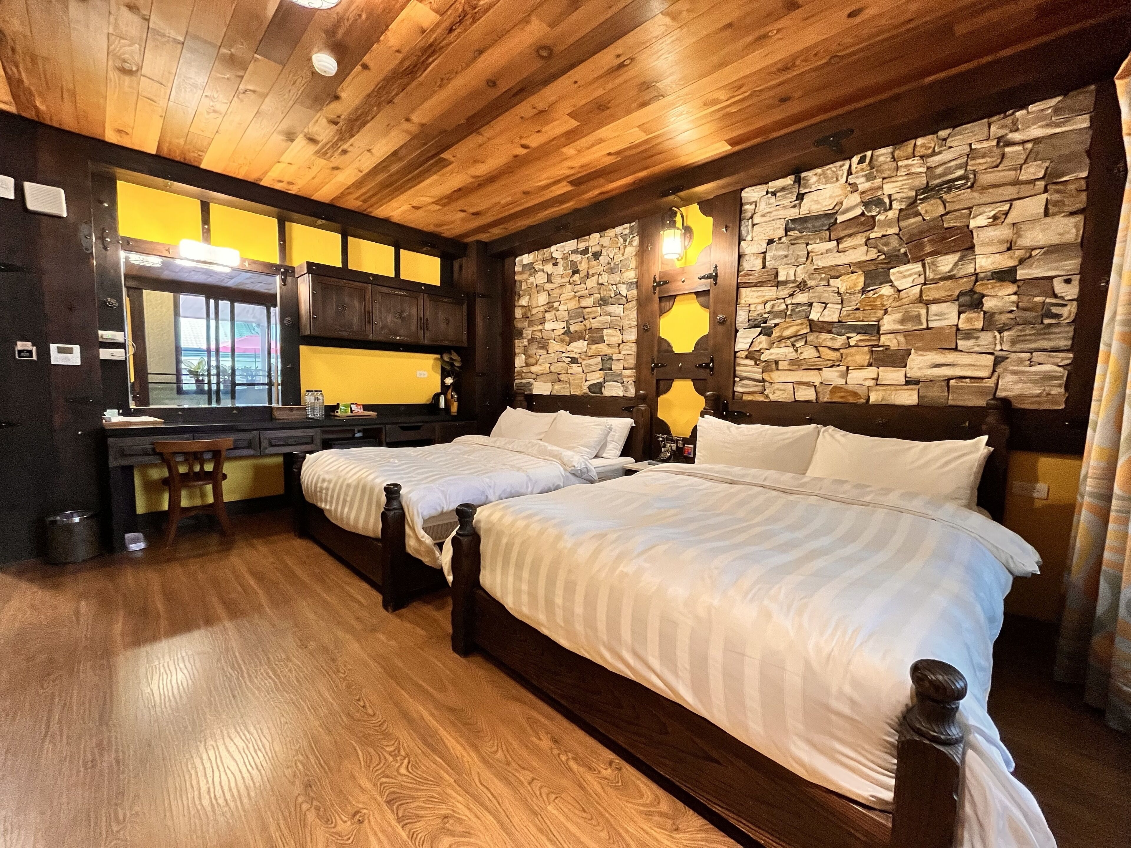 A hotel room with two beds, a wooden ceiling, a stone wall, and a desk with a mirror.