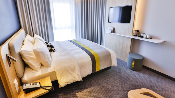 A hotel room with a bed, a bedside table with a telephone, a TV mounted on the wall, a small shelf with items, and a waste bin.