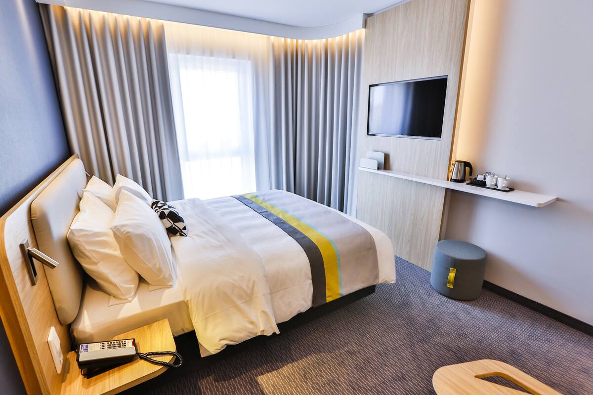 Une chambre d’hôtel comprenant un lit, une table de chevet avec un téléphone, un téléviseur fixé au mur, une petite étagère avec quelques objets et une poubelle.