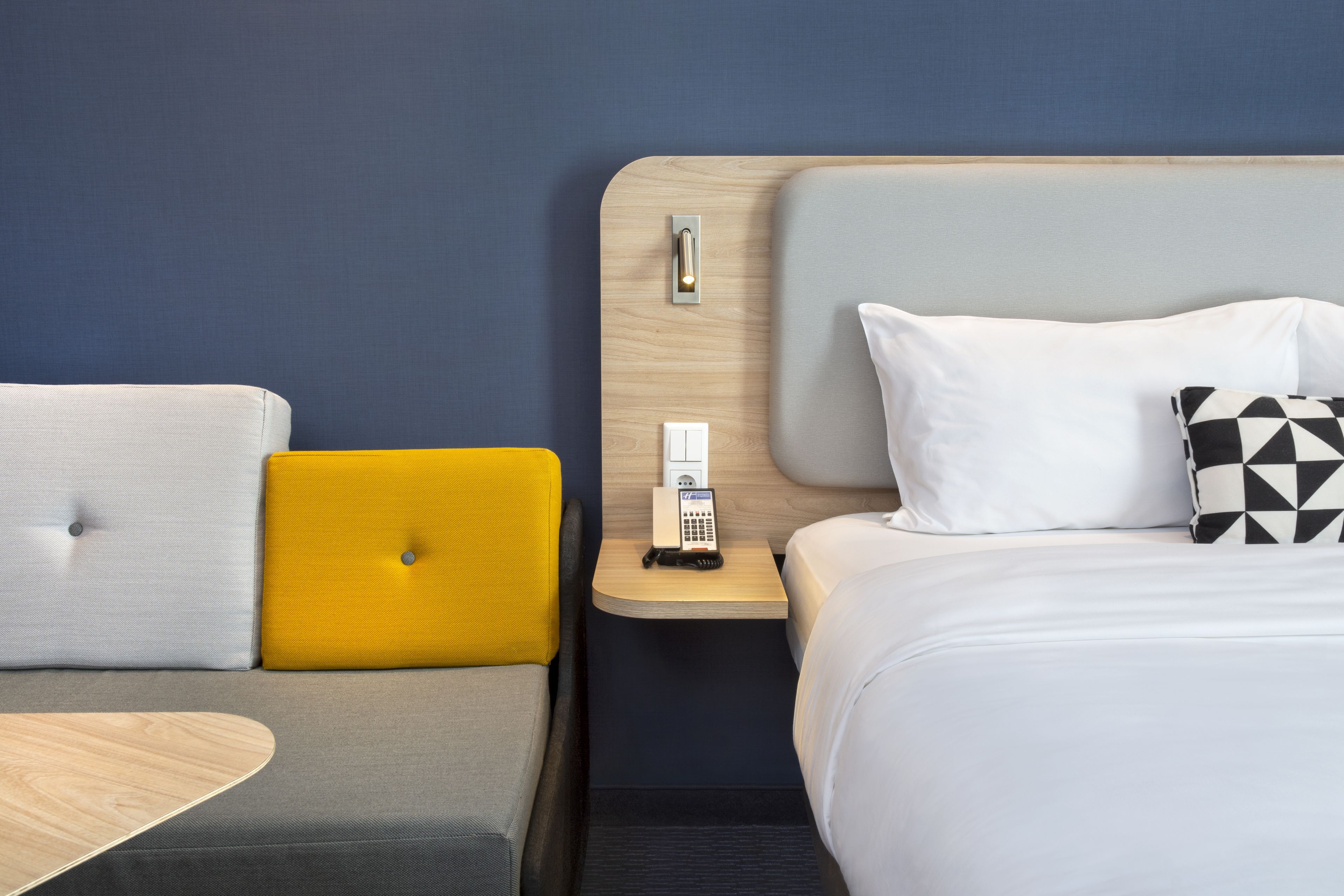 A hotel room with a bed, a bedside table with a phone, a nightstand, and a sofa with cushions.