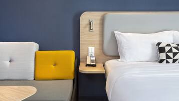 A hotel room with a bed, a bedside table with a phone, a nightstand, and a sofa with cushions.