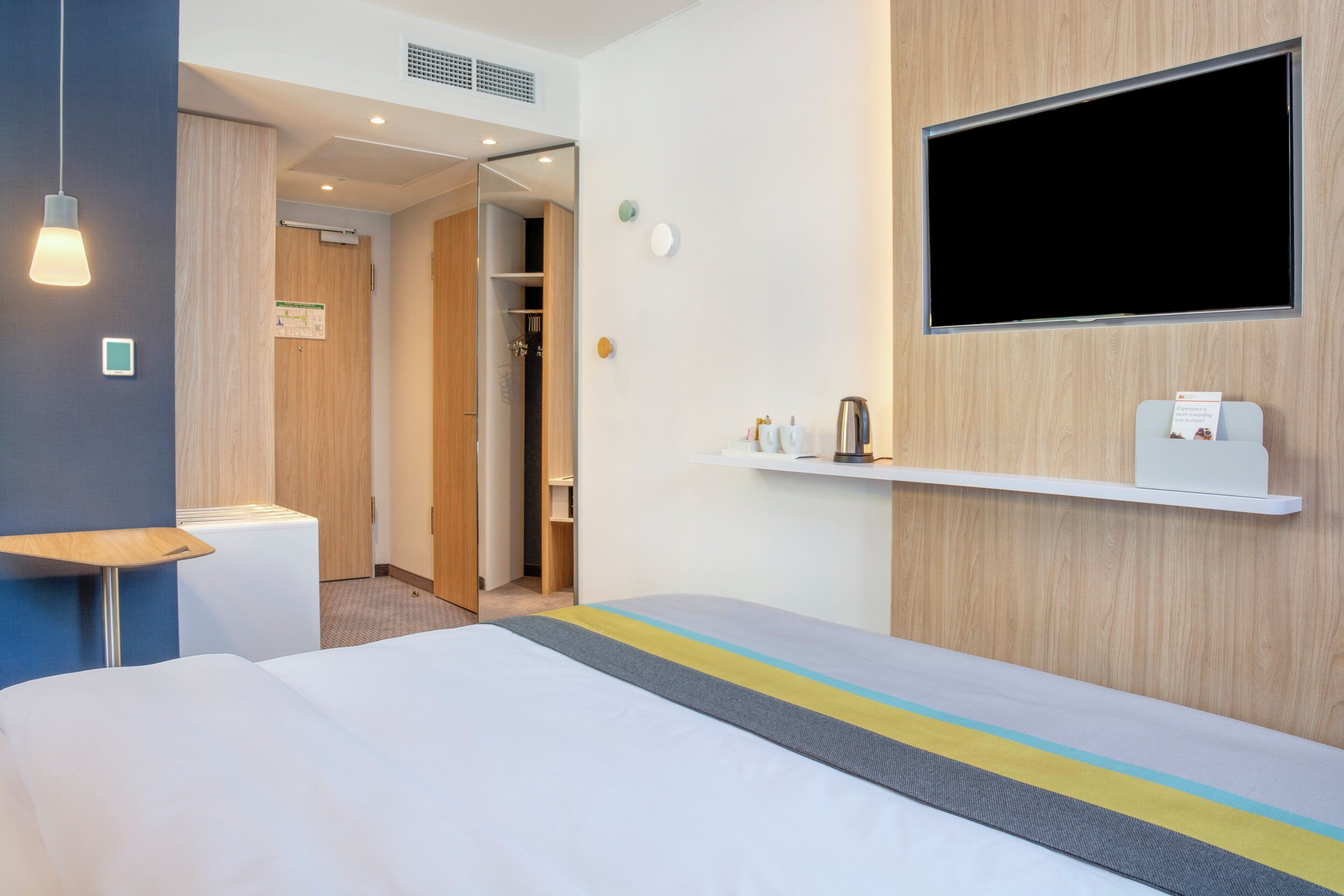 Une chambre d’hôtel avec un lit, une table de chevet, un téléviseur fixé au mur et une petite étagère avec quelques objets.