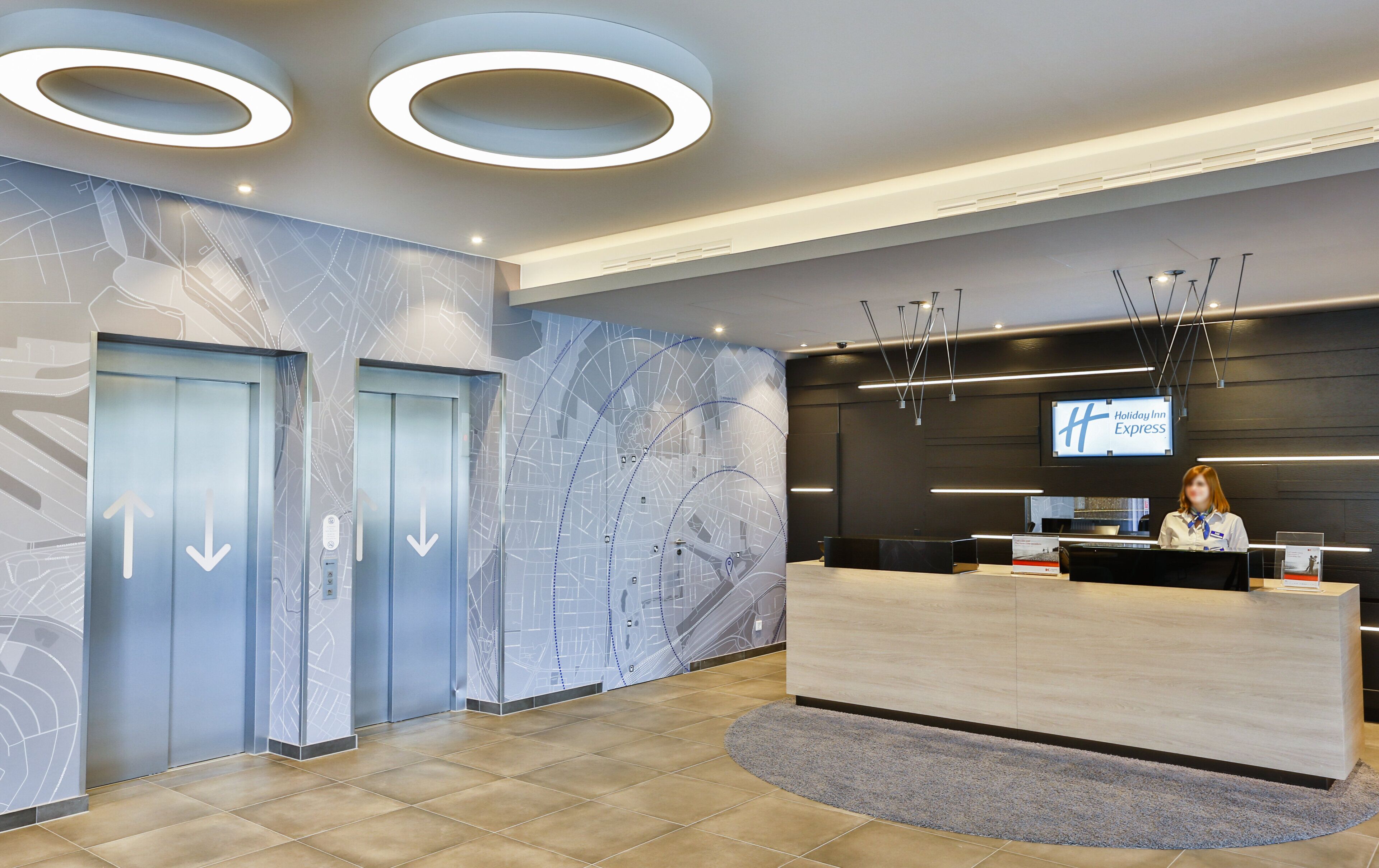 Zona de recepción de hotel con diseño moderno, que incluye un mostrador de recepción, una pantalla digital mostrando el nombre del hotel y dos ascensores con flechas direccionales.
