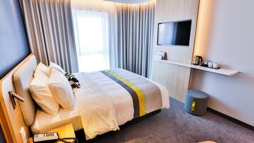 A hotel room with a bed, a TV mounted on the wall, a bedside table, a waste bin, and a small wooden stool.