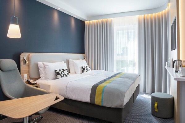 Une chambre d’hôtel moderne équipée d’un lit, d’un bureau, d’une chaise, d’une table de chevet et d’une fenêtre avec des rideaux.
