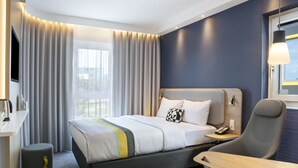 A modern hotel room with a bed, bedside table, chair, and a window with curtains.