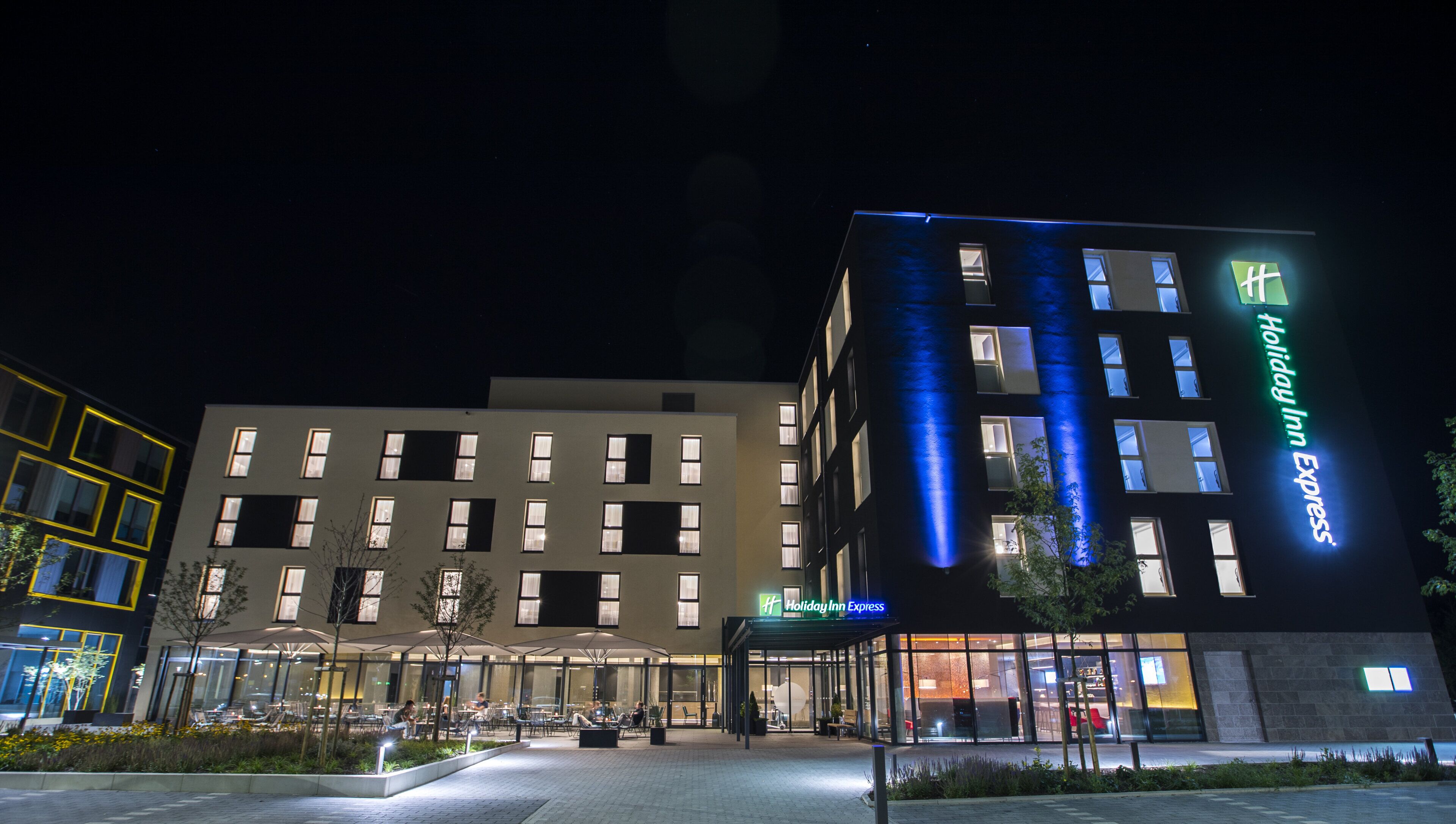 Un hotel Holiday Inn Express con diseño moderno, que presenta una entrada bien iluminada y un área de estar al aire libre.