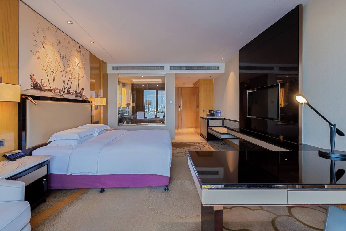 A modern hotel room with a large bed, a desk, a TV, and a view of the city through the window.