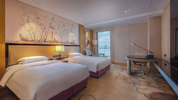 A hotel room with two beds, a desk, a chair, a lamp, and a large window with a city view.