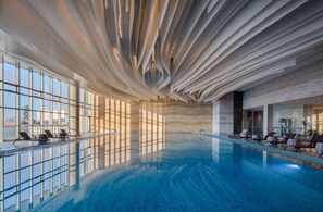 An indoor swimming pool with a modern ceiling design, large windows, and sun loungers.