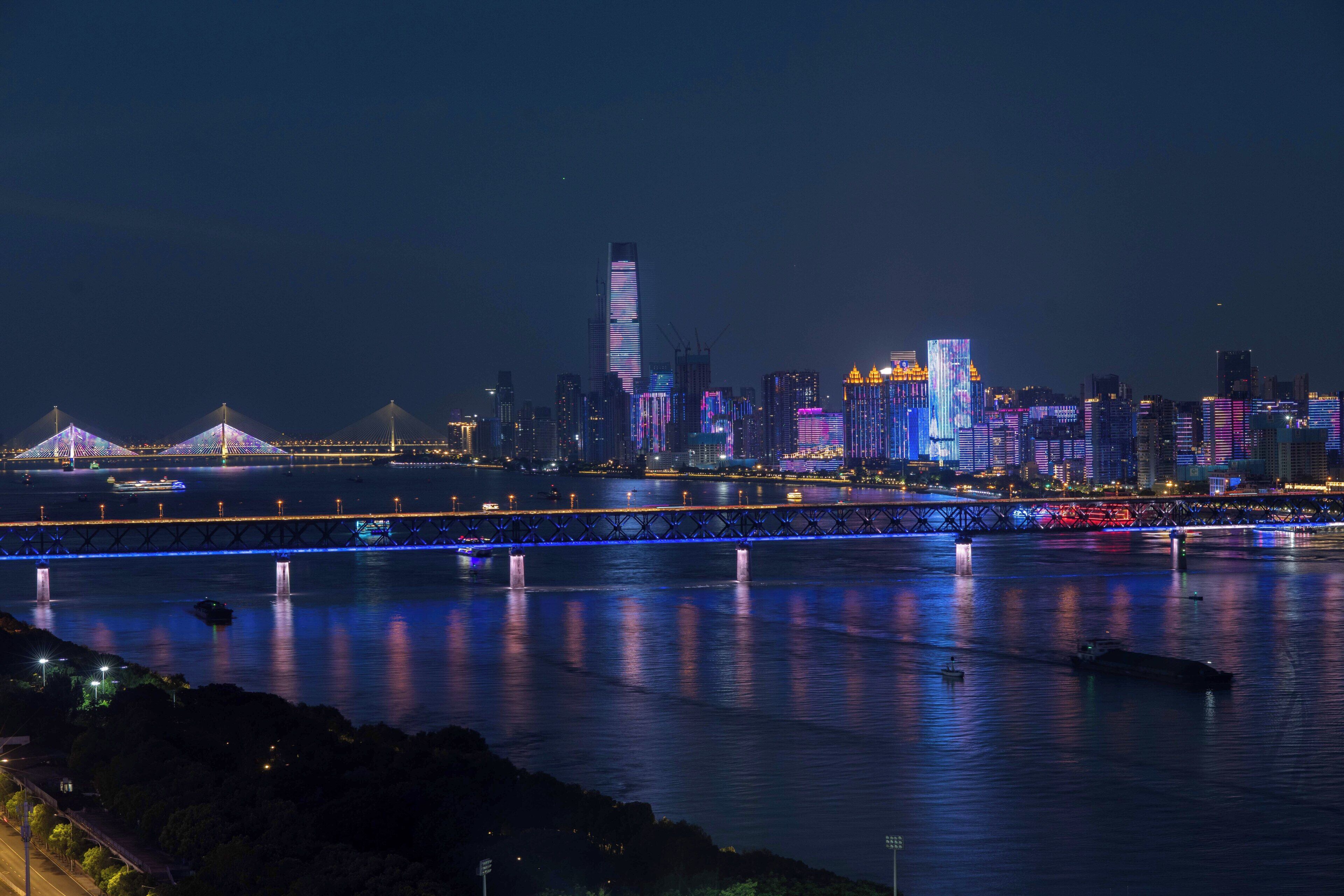 A nighttime cityscape with illuminated buildings and a bridge over a river.