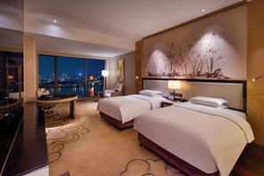 Hotelkamer met twee bedden, een bureau, een stoel, een groot raam met uitzicht op de stad en een decoratief object aan de muur.