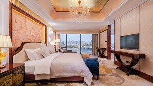 A spacious hotel room with a large bed, a television, a view of the city, and a chandelier.