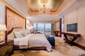 A spacious hotel room with a large bed, a television, a view of the city, and a chandelier.