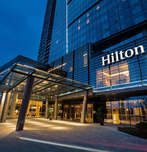 Hall de entrada de um hotel moderno com fachada de vidro e o nome 'Hilton' em destaque.