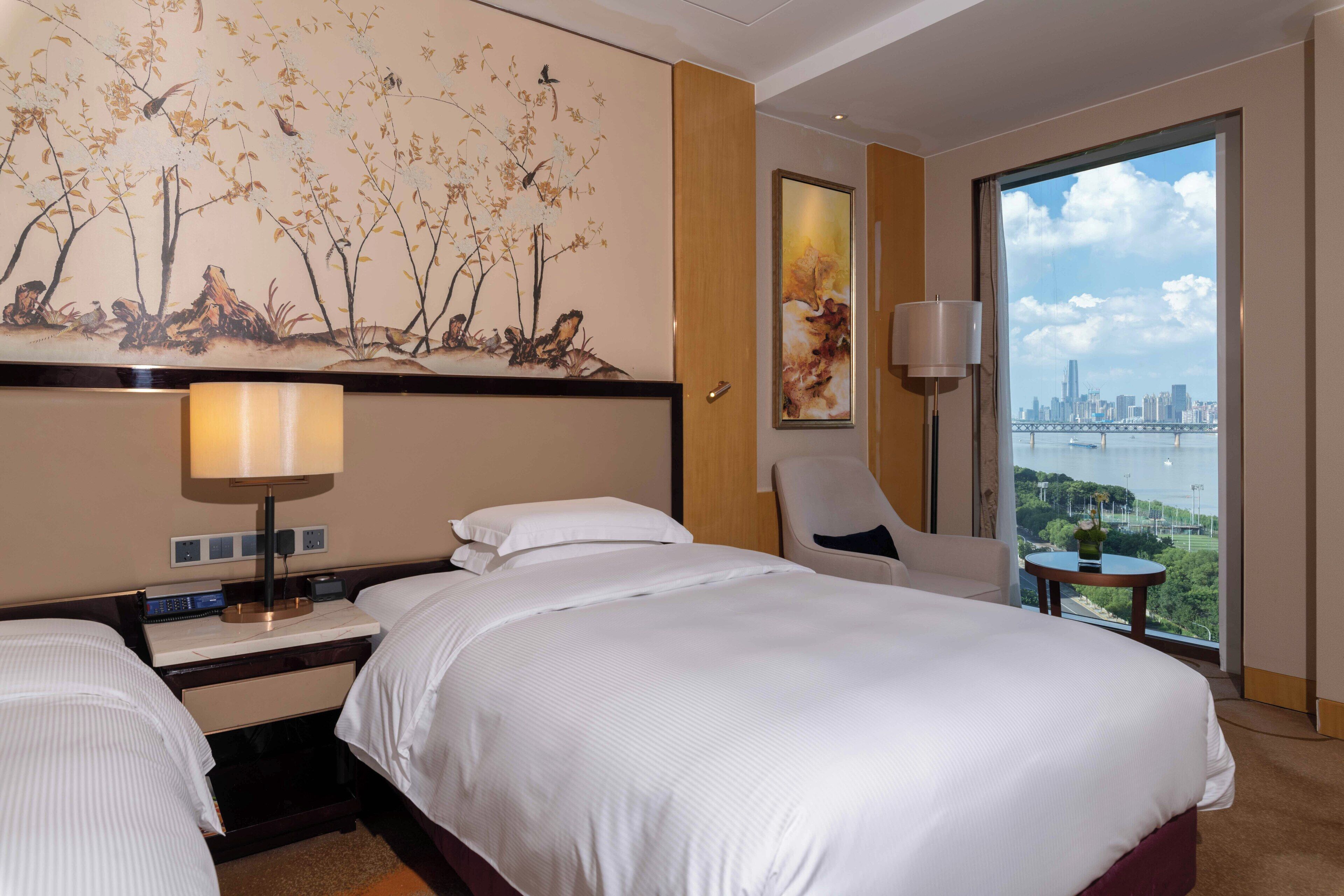Habitación de hotel con una cama grande, una silla, un escritorio con lámpara y vistas a la ciudad a través de un ventanal amplio.