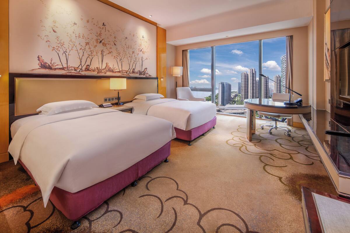A hotel room with two beds, a desk, a chair, a TV, and a large window with a city view.