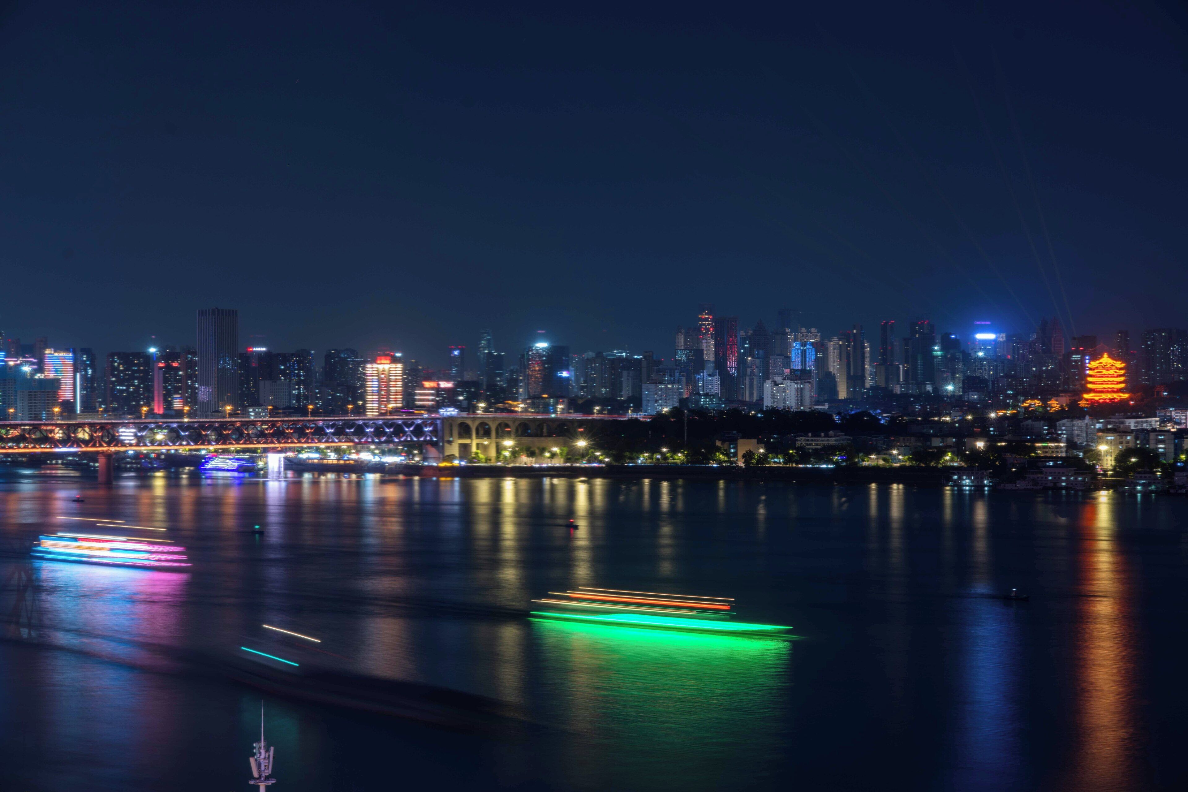 Une vue nocturne d’une ville, avec une rivière reflétant les lumières de la ville, un pont et une structure ressemblant à une pagode.