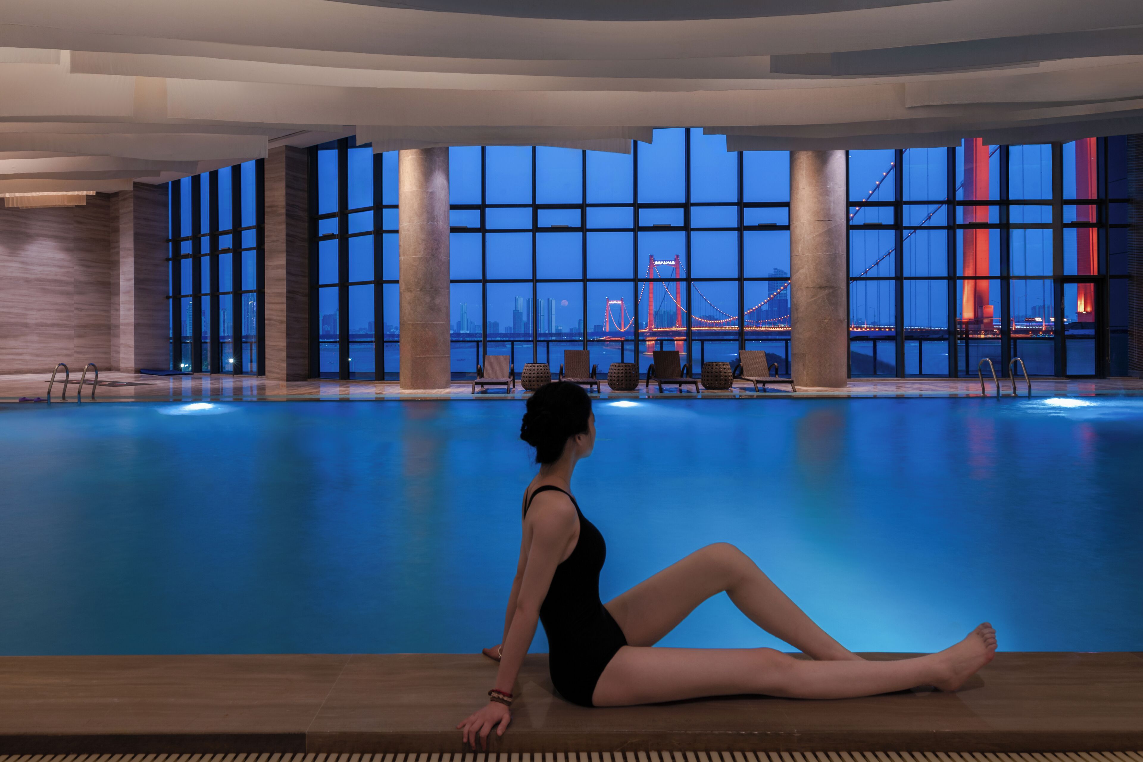 A woman sitting by a pool with a cityscape and bridge in the background.