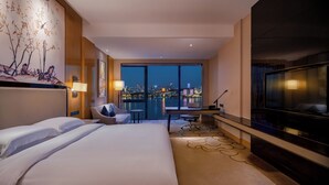 A hotel room with a large bed, a desk, a chair, a TV, and a view of the cityscape through large windows. - Hilton Wuhan Riverside (Wuhan)